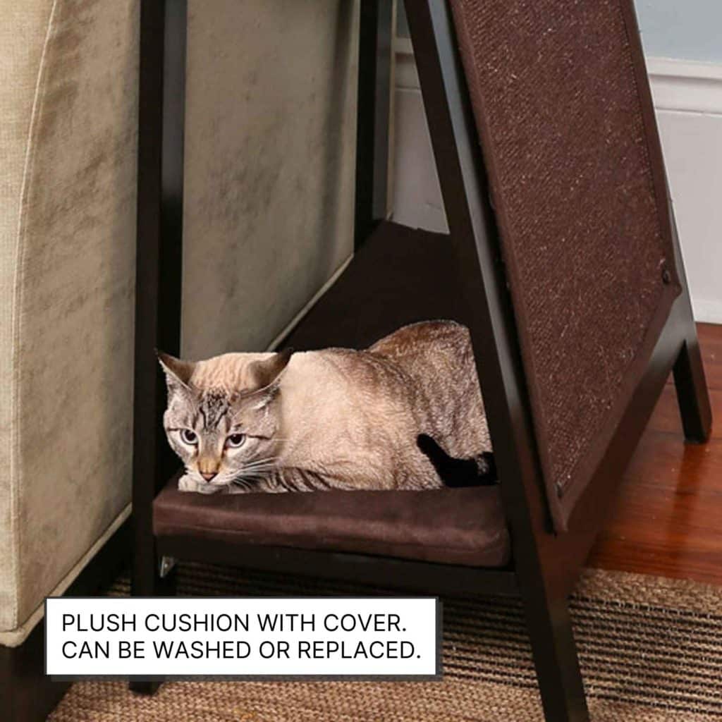 A cat relaxes on an A-frame wooden cat bed with a brown plush cushion under a table next to a beige couch.