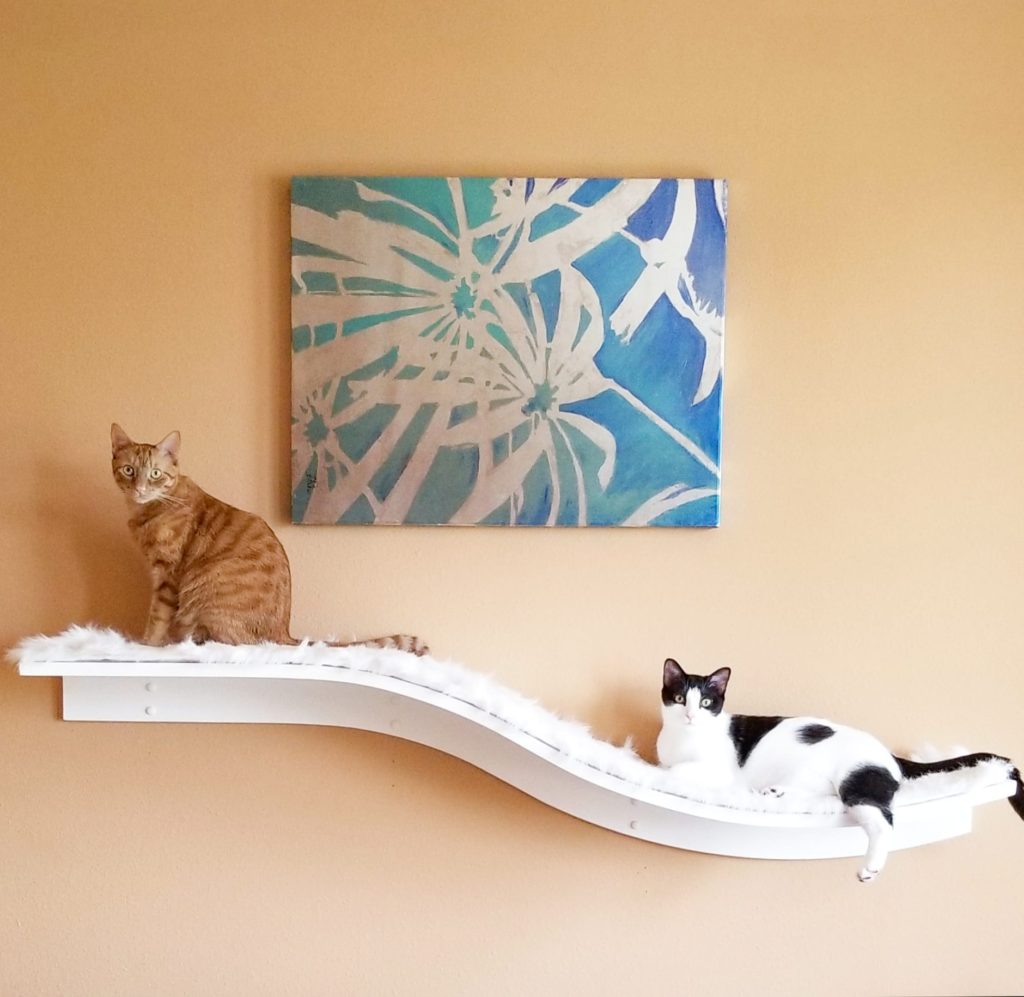 Two cats lounging on a white, fluffy Lotus Branch Cat Shelf beneath a blue abstract painting on a beige wall.