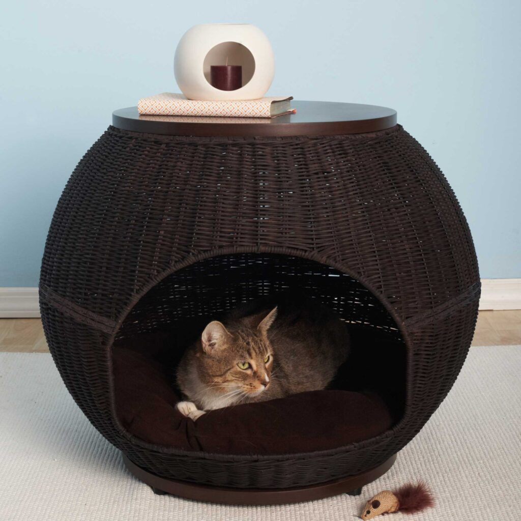 A tabby cat rests inside a round wicker bed that doubles as a side table holding a toy mouse, books, and a vase.