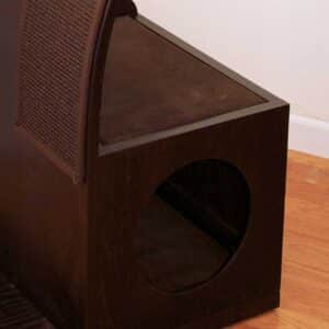 Dark wooden pet house with circular entrance, angled mesh panel, and cushion on hardwood floor by a white wall.