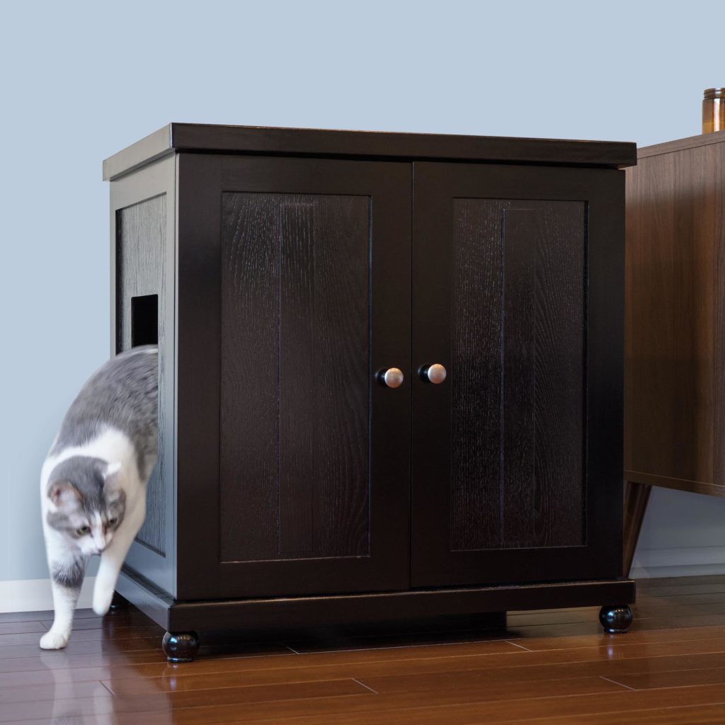 Gray and white cat steps out of a dark wooden cabinet with side entrance, on a wooden floor next to another furniture piece.