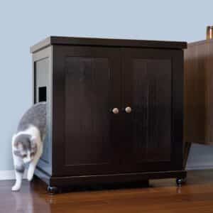 Gray and white cat steps out of a dark wooden cabinet with side entrance, on a wooden floor next to another furniture piece.