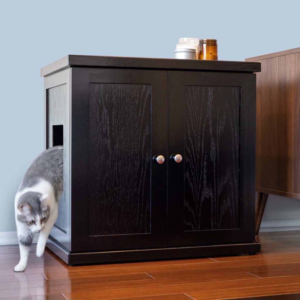Gray and white cat steps out of a black litter box cabinet with side opening, next to furniture on wood floor.