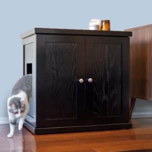 Gray and white cat steps out of a black litter box cabinet with side opening, next to furniture on wood floor.