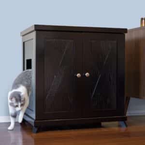 A gray and white cat walks out of a dark wood litter box cabinet on a wooden floor next to other furniture.