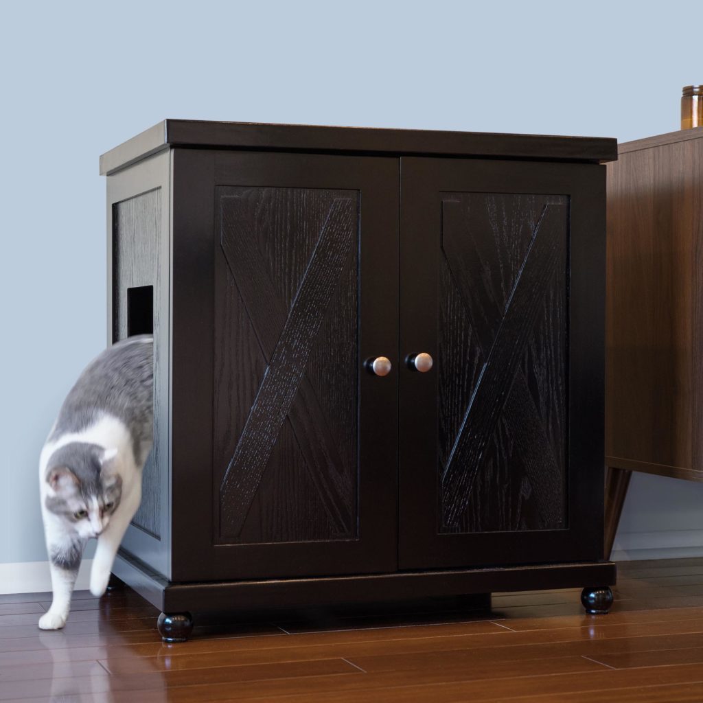 Gray and white cat stepping out of a dark wood litter box cabinet on hardwood floor next to other furniture.