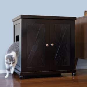Gray and white cat stepping out of a dark wood litter box cabinet on hardwood floor next to other furniture.