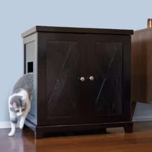 Gray and white cat stepping from a dark wood side-entry litter box on hardwood floor next to another furniture piece.