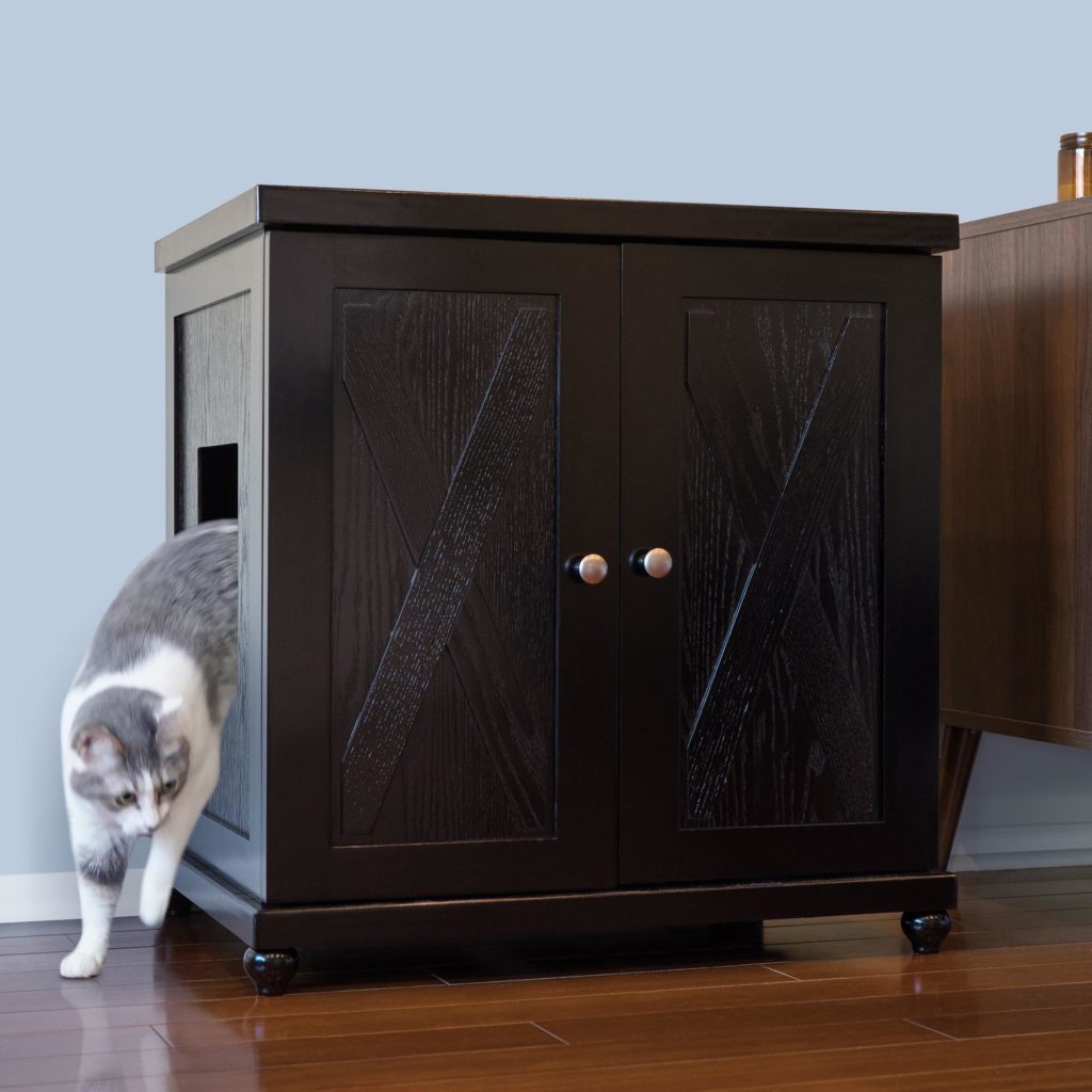 Gray and white cat stepping out of a dark wooden double-door litter box cabinet on a hardwood floor.