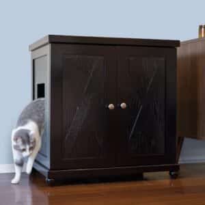 Gray and white cat stepping out of a dark wooden double-door litter box cabinet on a hardwood floor.