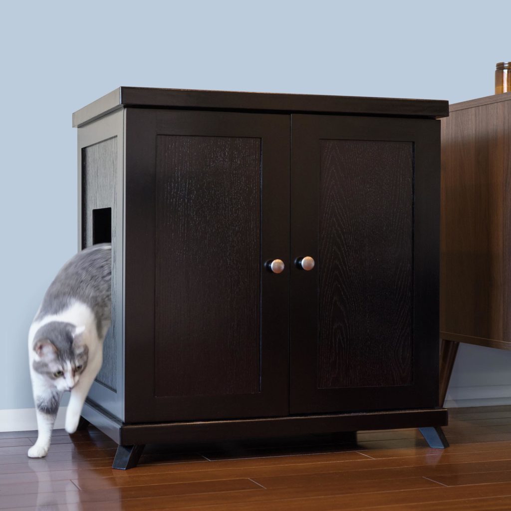 Gray and white cat exits a dark wood litter box with side opening on hardwood floor, next to a light blue wall.