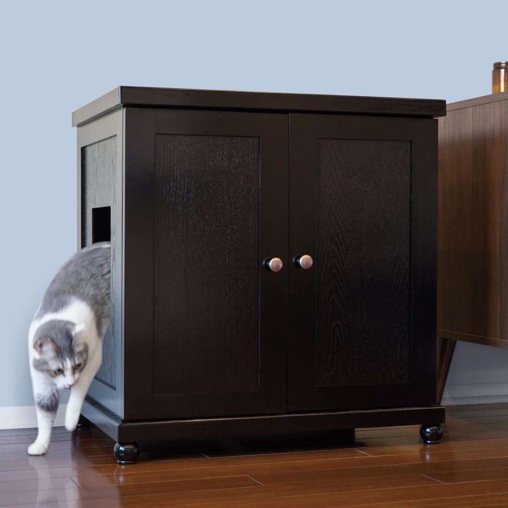 A gray and white cat steps out of a round dark wood litter box cabinet on a wooden floor by a light blue wall.