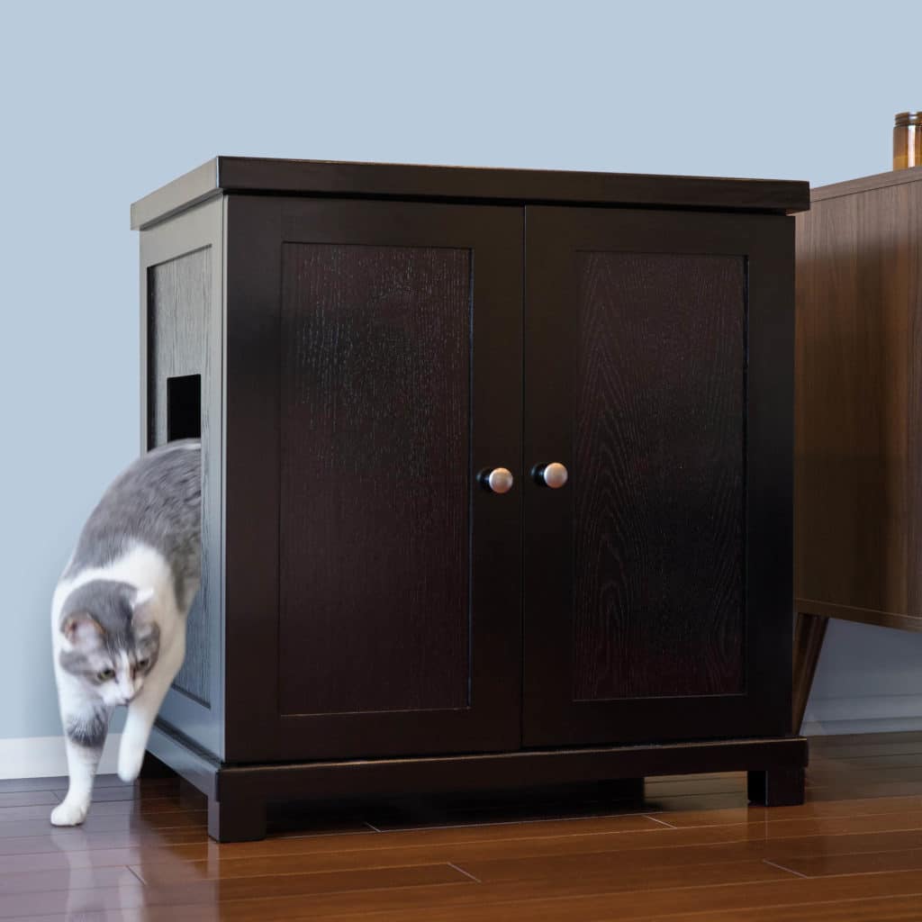 Gray and white cat steps out of a dark wood litter box with side opening on wooden floor near a light blue wall.