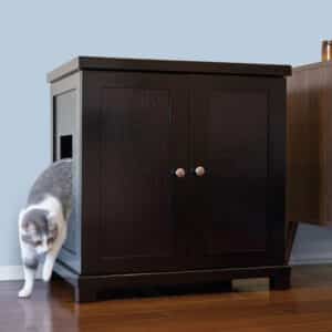 Gray and white cat steps out of a dark wood litter box with side opening on wooden floor near a light blue wall.