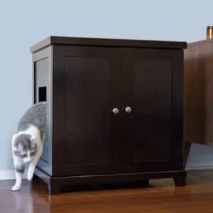 Gray and white cat stepping out of a side-opening litter box cabinet on hardwood floor beside another furniture piece.