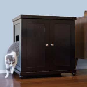 A gray and white cat steps out of a dark wood-style litter box on a hardwood floor near a light blue wall.