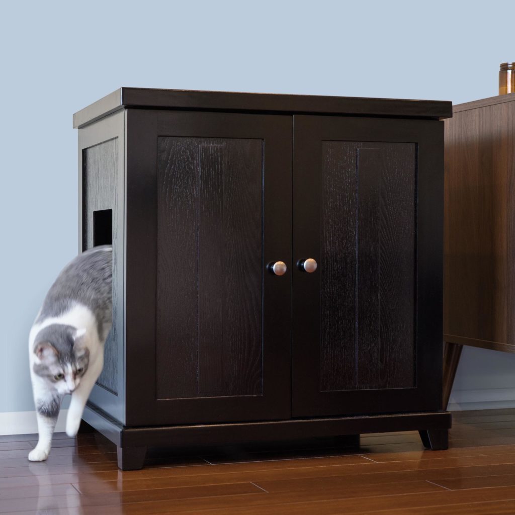 Gray and white cat exiting side opening of dark wood litter box cabinet near a light blue wall on hardwood floor.