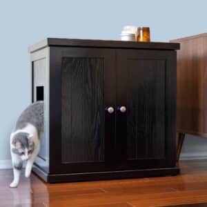 Gray and white cat leaves a black deluxe litter box with side opening; two cat litter containers rest on top of the cabinet.