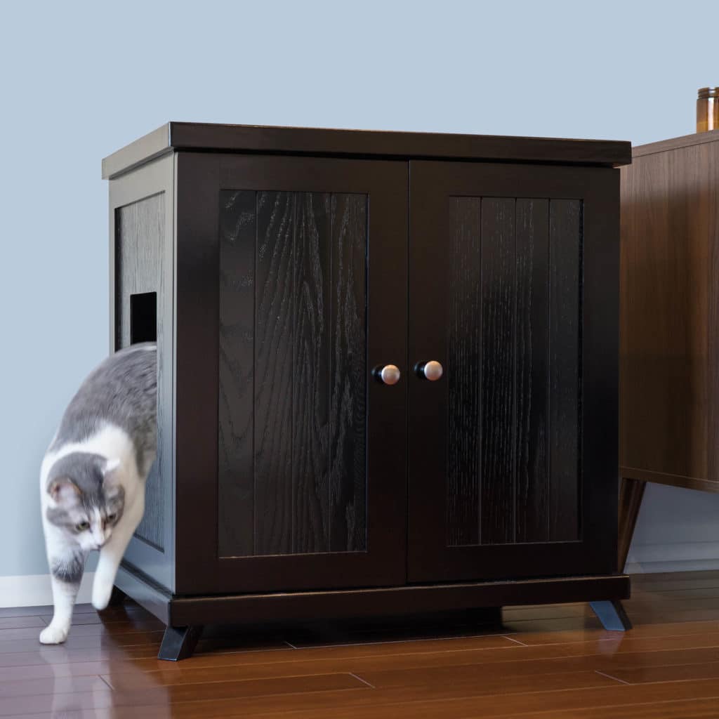 Gray and white cat steps out of a dark wooden litter box with side opening on hardwood floor next to furniture.