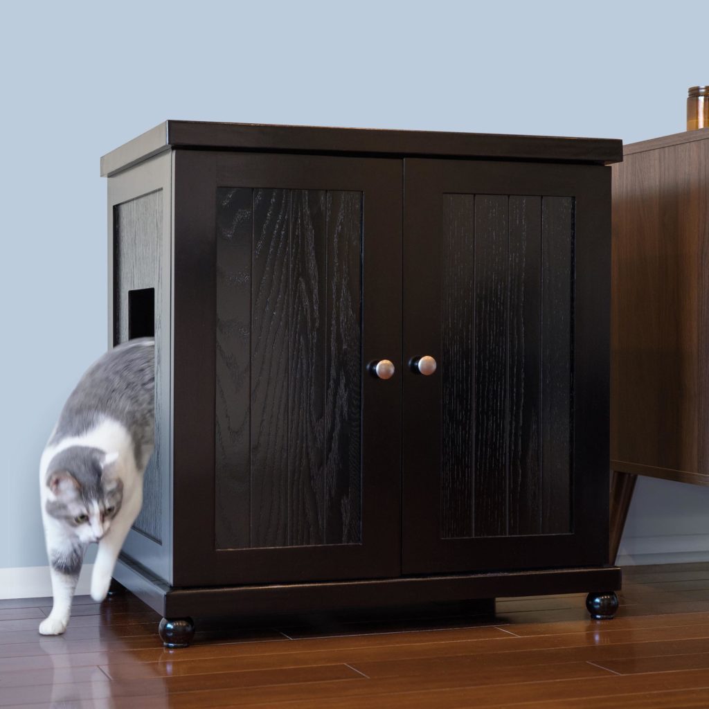 Gray and white cat exits a dark wood cat box with side opening on hardwood floor next to another furniture piece.