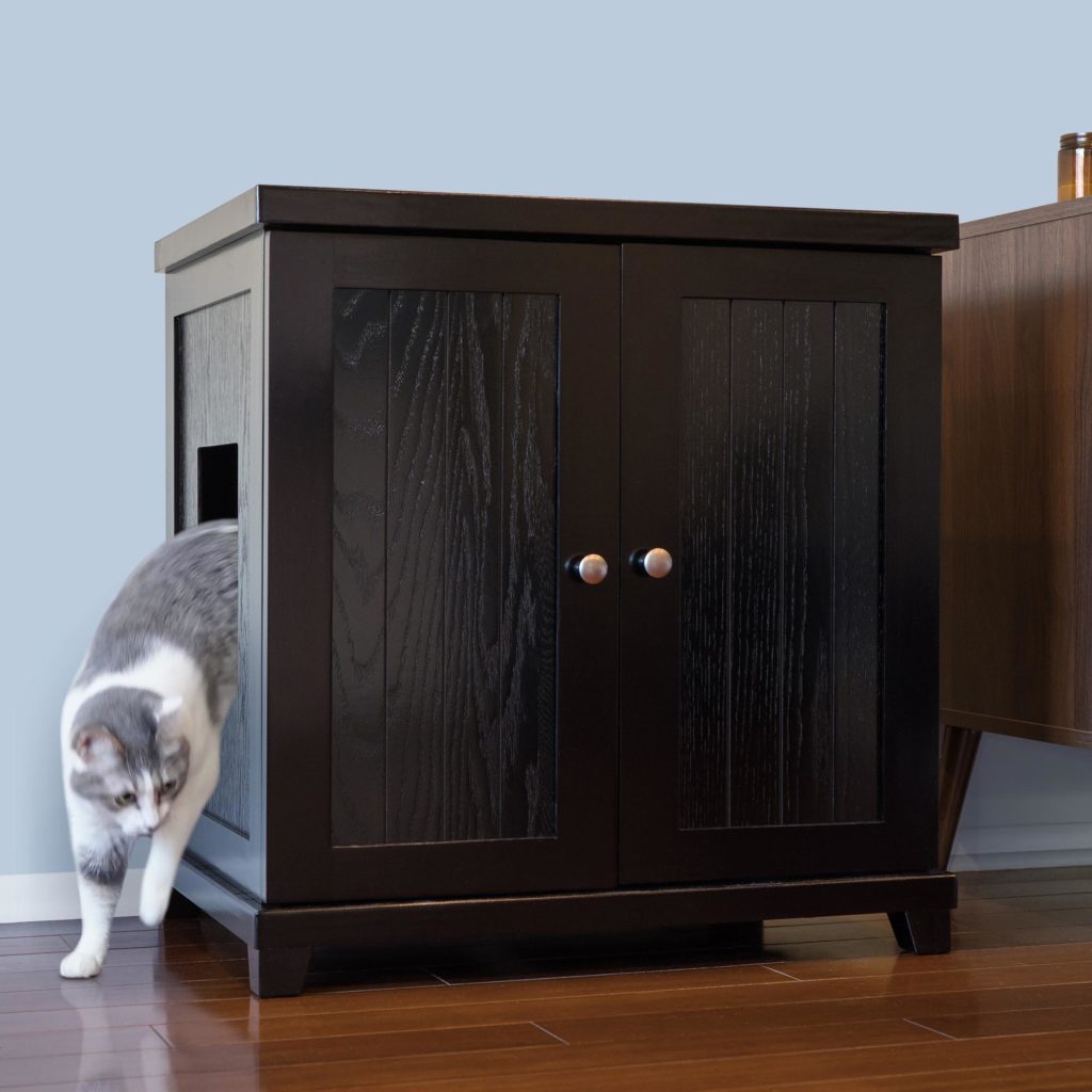 A gray and white cat steps out of a dark wooden cabinet with a side opening on a hardwood floor, hiding a litter box inside.