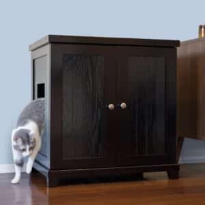 A gray and white cat steps out of a dark wooden cabinet with a side opening on a hardwood floor, hiding a litter box inside.