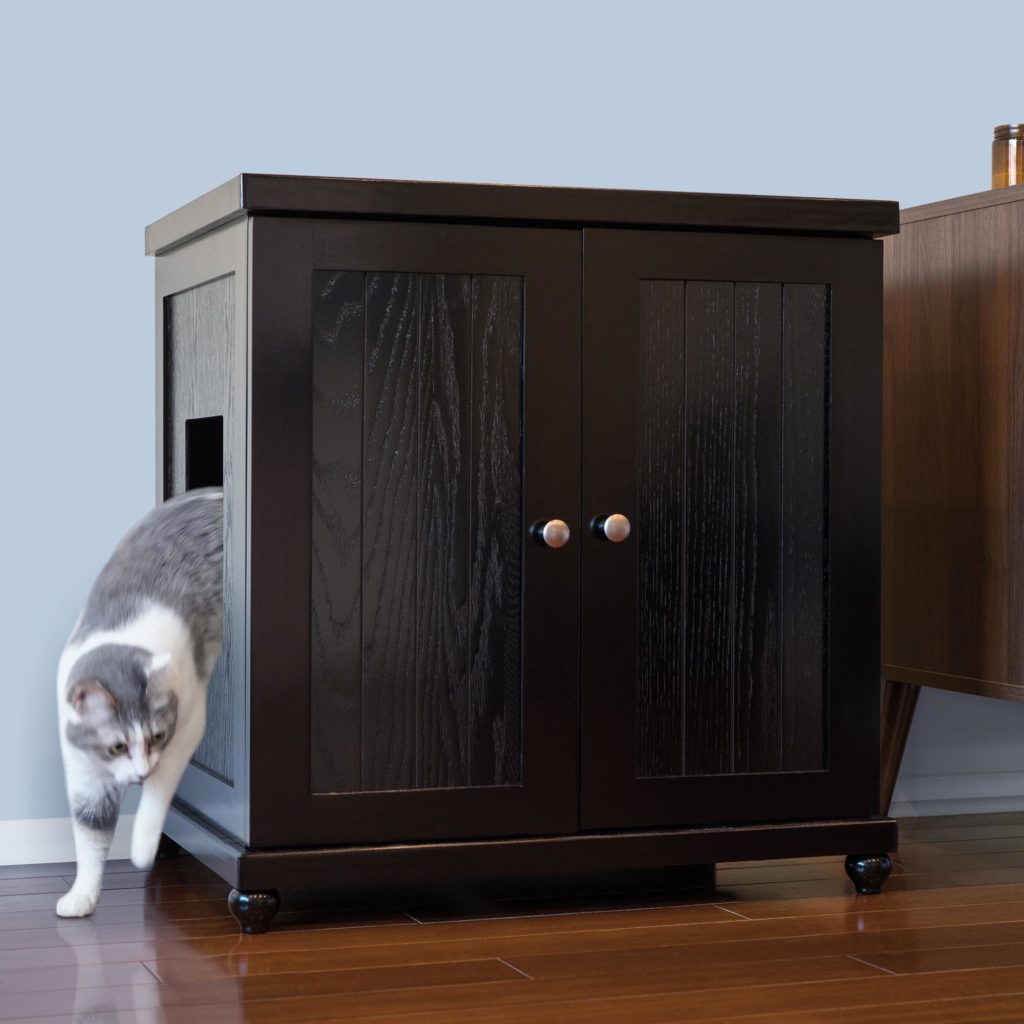 A cat steps out of a black wooden litter box with a side opening, on hardwood floor beside another piece of furniture.