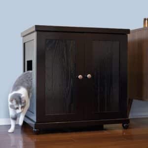 A cat steps out of a black wooden litter box with a side opening, on hardwood floor beside another piece of furniture.