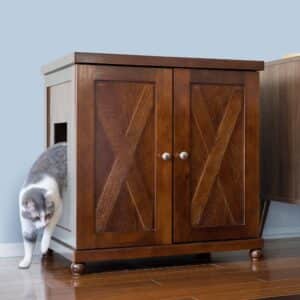 Gray and white cat exits a side opening of a round litter box cabinet with two front doors on hardwood floor.