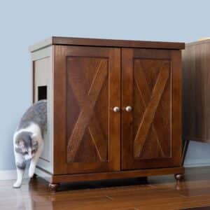 Gray and white cat exits a side opening in a double-door litter box cabinet on hardwood floor near a light blue wall.