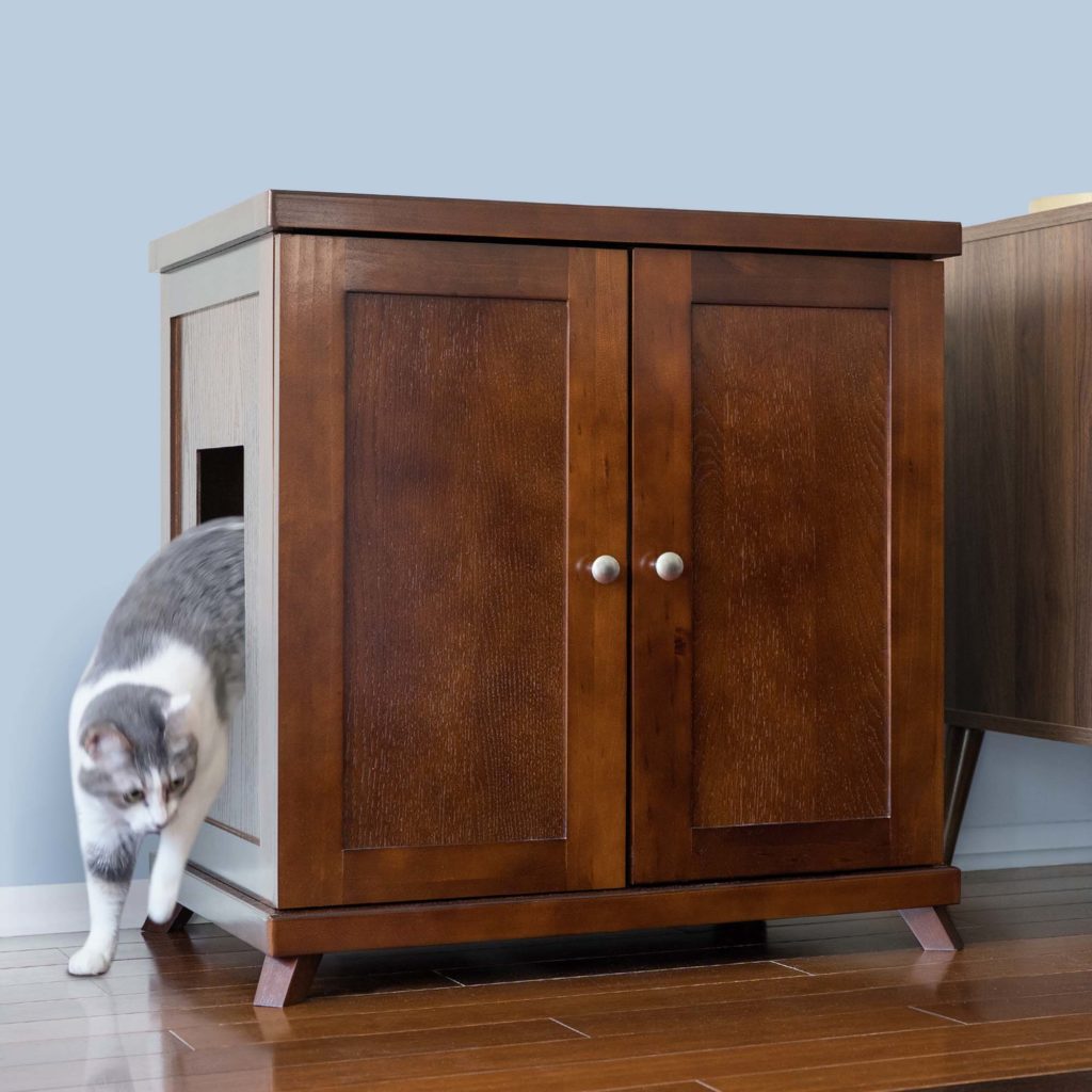 Gray and white cat leaving a cabinet-style litter box with side opening on wooden floor near light blue wall.