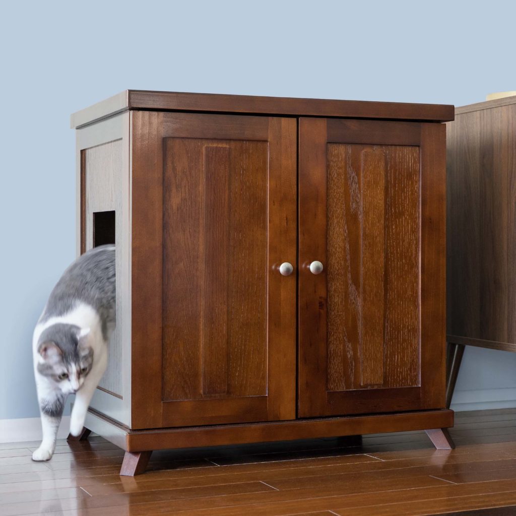 Gray and white cat steps from the side opening of a wooden litter box cabinet with double doors on hardwood floor.