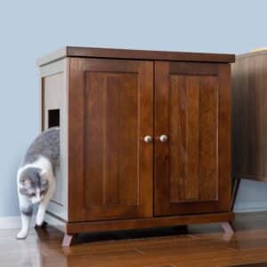 Gray and white cat steps from the side opening of a wooden litter box cabinet with double doors on hardwood floor.