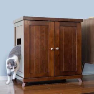 Gray and white cat stepping out of a dark wood litter box cabinet with side opening on wooden floor by a light blue wall.