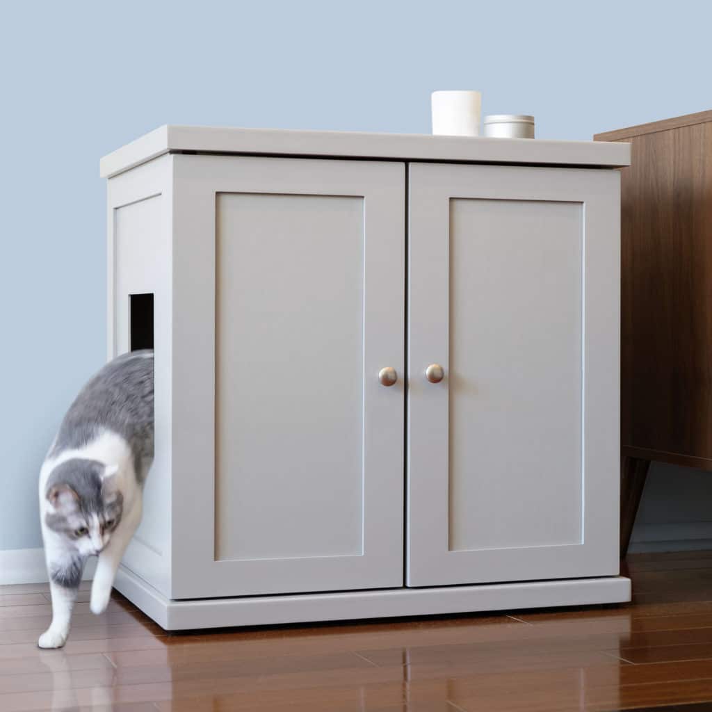 Gray and white cat exiting a small door on a light gray litter box cabinet on wooden floor beside brown furniture.