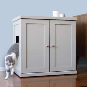 Gray and white cat exiting a small door on a light gray litter box cabinet on wooden floor beside brown furniture.