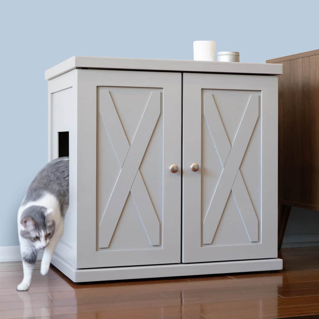 Gray and white cat exits the side of a paneled litter box cabinet on wooden floor beside brown furniture.