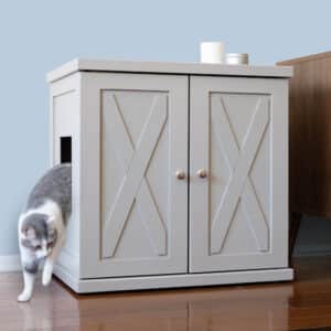 Gray and white cat exits the side of a paneled litter box cabinet on wooden floor beside brown furniture.