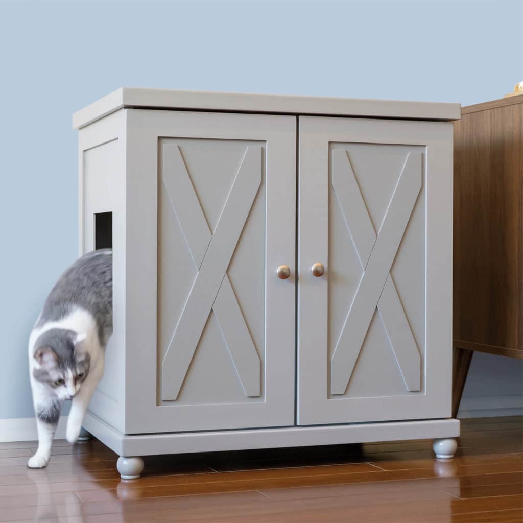 Cat walking out of gray litter box cabinet with X-design doors, on wooden floor beside brown furniture.