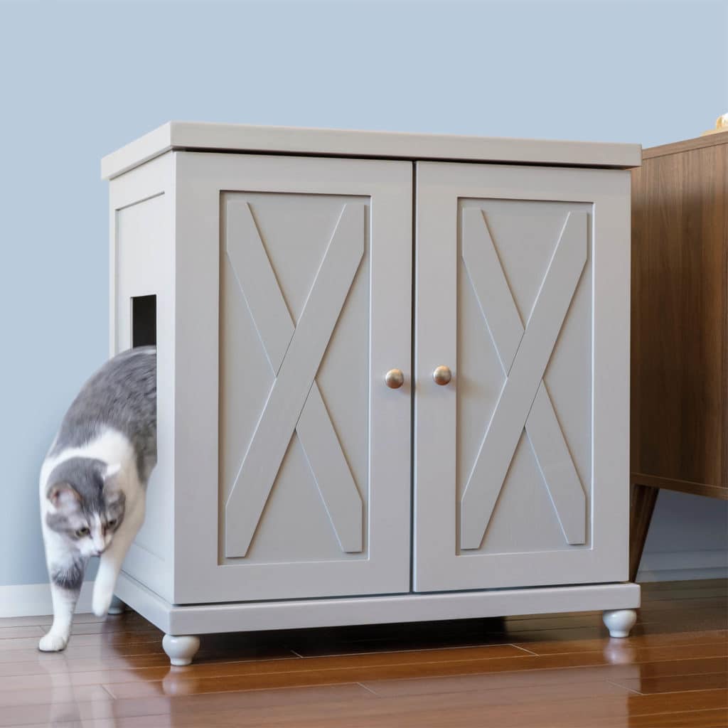 Gray and white cat leaves a double-door litter box cabinet on wood floor next to a light blue wall.