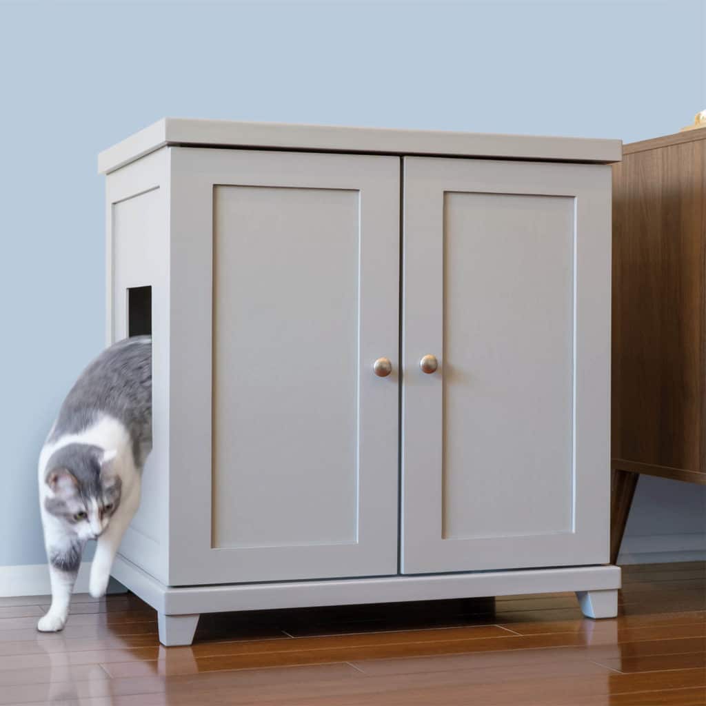 Gray and white cat exits a side-opening litter box cabinet on a wooden floor beside another piece of furniture.