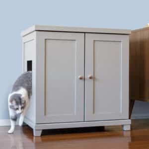 Gray and white cat exits a side-opening litter box cabinet on a wooden floor beside another piece of furniture.