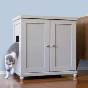 Gray and white cat exiting a side opening of a double-door litter box cabinet on a wooden floor.