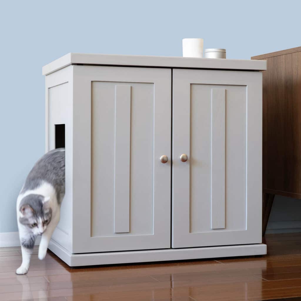 A gray and white cat leaves a cabinet litter box with a side opening on a wooden floor next to brown furniture.