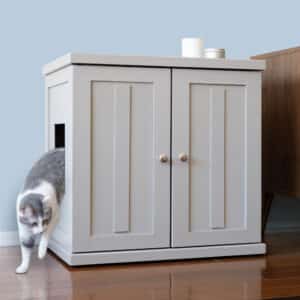 A gray and white cat leaves a cabinet litter box with a side opening on a wooden floor next to brown furniture.