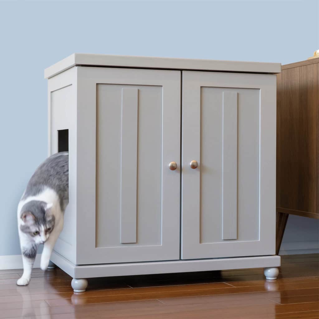 A gray and white cat steps out of a light gray litter box cabinet with a side opening, set on a wooden floor.
