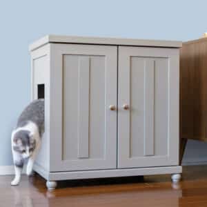 A gray and white cat steps out of a light gray litter box cabinet with a side opening, set on a wooden floor.