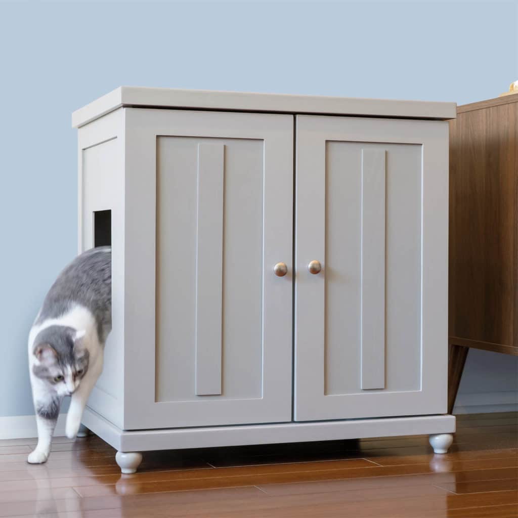 A gray and white cat exits a side door of a light gray wooden litter box cabinet on a wooden floor.