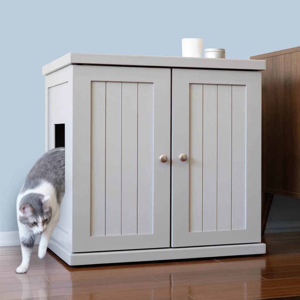 Gray and white cat exits a small side door of a white litter box cabinet with double doors on a wooden floor by brown furniture.
