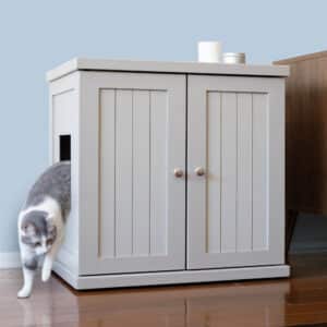 Gray and white cat exits a small side door of a white litter box cabinet with double doors on a wooden floor by brown furniture.